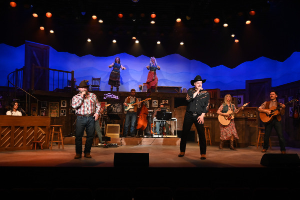 Two lead vocalists sing at centre stage during a lively number in Nashville Music City. Wearing cowboy hats and western-inspired attire, they perform with microphones while musicians and additional singers accompany them from the surrounding set and elevated platform. Warm stage lighting and dynamic poses capture the excitement of a live country music showcase.