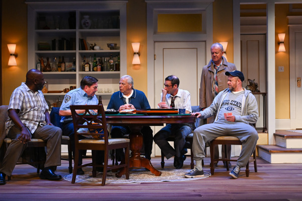 Six performers gather around a dining table in a warmly lit apartment set. Several sit with playing cards and drinks while one performer in a police uniform leans toward another across the table, and a performer in a jacket stands behind the group observing the conversation.