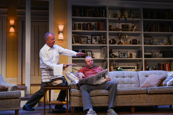 One performer relaxes on a sofa reading a newspaper while another approaches from behind carrying a plate of food, a towel draped over one arm, and utensils held out toward him. Bookshelves filled with framed photographs, trophies, books, and football memorabilia cover the wall behind them.
