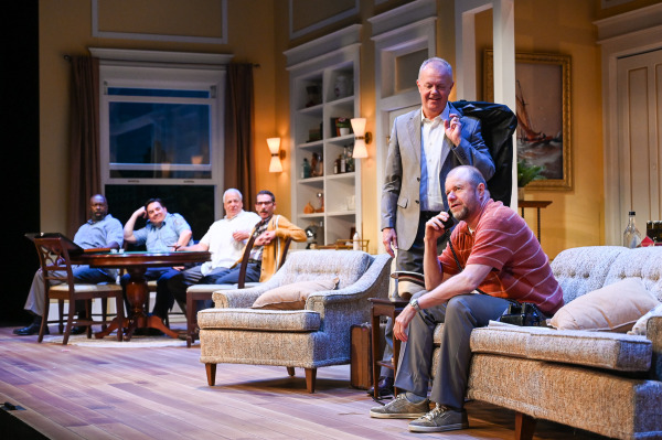 A performer in a red striped polo sits on a sofa speaking into a telephone while another stands behind him in a suit with a garment bag slung over his shoulder. Four other performers sit around a dining table in the background, watching the conversation from across the apartment.