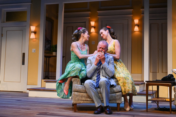 A performer in a light grey suit sits in an armchair holding a handkerchief to his face as two performers in colourful floral dresses lean close on either side, resting reassuring hands on his shoulders. The warm apartment interior and a black telephone on a nearby side table frame the scene.