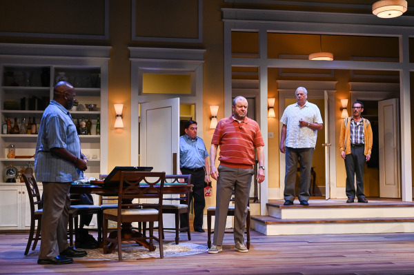 Five performers stand throughout a warmly lit living room and dining area during a tense moment. One stands beside the dining table at left, another in a red striped polo stands centre stage with his eyes closed, while three others watch from the doorway and raised entryway behind him.
