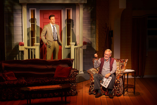 In a warmly lit scene from Drayton Entertainment’s Over the River and Through the Woods at King’s Wharf Theatre, a younger performer in a checked blazer and tie stands near a red front door with hands in pockets, watching as an older performer laughs and gestures from a floral armchair while holding a newspaper. A patterned sofa, coffee table, wood flooring, and detailed front porch complete the family home set.