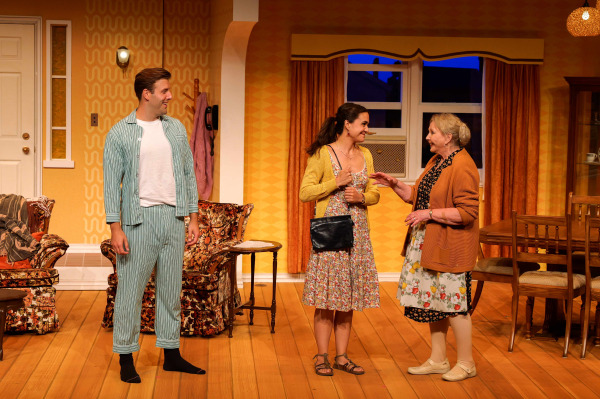 In a warmly lit scene from Drayton Entertainment’s Over the River and Through the Woods at King’s Wharf Theatre, a younger performer in striped pyjamas smiles as a woman in a floral dress and yellow cardigan speaks with an older performer wearing a flowered apron and orange cardigan. They stand together in the retro living and dining room set.