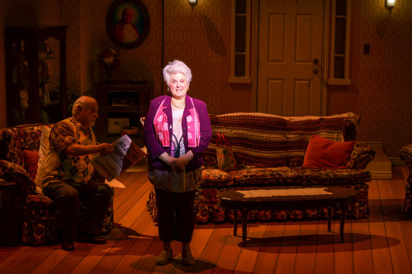 In a spotlighted scene from Drayton Entertainment’s Over the River and Through the Woods at King’s Wharf Theatre, an older performer in a purple jacket and pink scarf stands centre stage addressing the audience while another sits nearby reading a newspaper. Floral furniture, a crocheted sofa throw, patterned wallpaper, and warm amber lighting complete the family living room set.