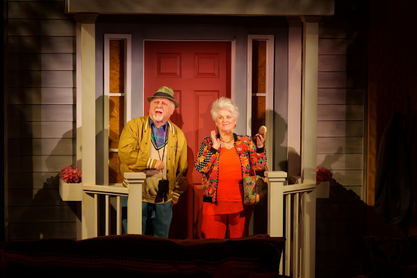 Two older performers stand on the front porch of a family home in Drayton Entertainment’s Over the River and Through the Woods at King’s Wharf Theatre. One wears a brimmed hat and mustard jacket, while the other, dressed in bright red with a colourful floral jacket, smiles and holds up a small bread roll beside the red front door.