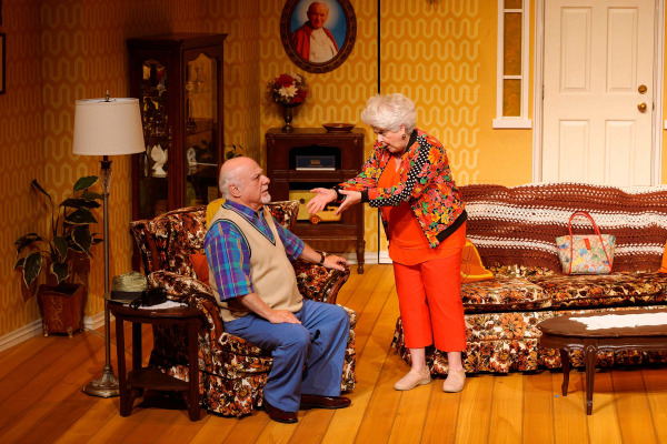 In a warmly lit living room scene from Drayton Entertainment’s Over the River and Through the Woods at King’s Wharf Theatre, an older performer sits in a floral armchair while another stands before him, gesturing emphatically with both hands. Retro patterned wallpaper, matching floral furniture, a display cabinet, lamp, and framed portrait complete the detailed family home set.