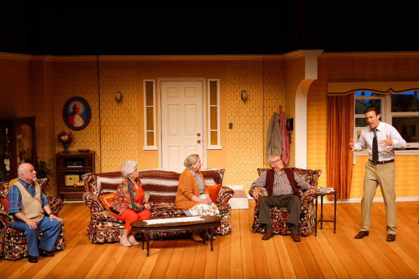 In a scene from Drayton Entertainment’s Over the River and Through the Woods at King’s Wharf Theatre, four older performers sit across a retro living room while a younger performer stands at the side, speaking animatedly with his hands raised. Floral furniture, patterned wallpaper, framed décor, and warm lighting create the atmosphere of a welcoming family home.