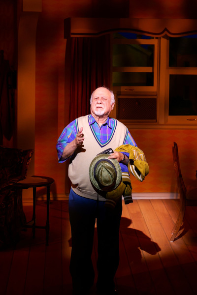 In a spotlighted scene from Drayton Entertainment’s Over the River and Through the Woods at King’s Wharf Theatre, an older performer gestures while holding a brimmed hat, mustard jacket, and small camera. He stands alone in the warmly lit living room set, dressed in a colourful plaid shirt, beige sweater vest, and blue trousers.