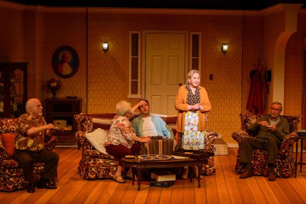 In a living room scene from Drayton Entertainment’s Over the River and Through the Woods at King’s Wharf Theatre, a younger performer reclines on the floral sofa with a hand to his forehead as an older performer leans toward him. Three others gather nearby with snacks while a board game and open box sit on the coffee table, creating a lively family gathering in the warmly lit home set.
