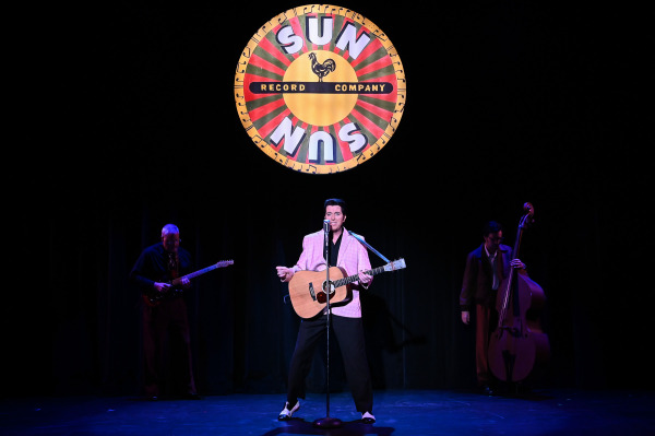 A performer portraying Elvis Presley stands at a vintage microphone with an acoustic guitar beneath a large 