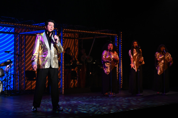 A performer in a gold jacket sings into a handheld microphone during Return to Grace at Drayton Entertainment. Three backing vocalists in matching gold tops perform behind him while the band plays beneath warm concert lighting.