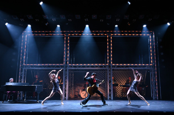 A performer playing guitar strikes a dynamic pose centre stage in Return to Grace at Drayton Entertainment while two dancers in sequined costumes mirror each other on either side beneath bright white spotlights. The live band performs behind a multi-level illuminated set.