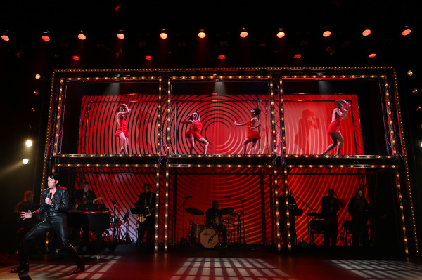 A performer portraying Elvis Presley sings at stage left in Return to Grace at Drayton Entertainment while four dancers perform on elevated platforms behind him. The stage is bathed in red lighting with bold circular graphic backdrops and a live band visible in the background