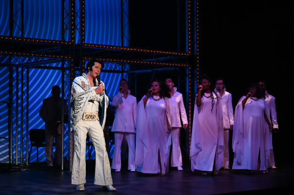 A performer portraying Elvis Presley, dressed in an iconic white jumpsuit, sings into a microphone during Return to Grace at Drayton Entertainment. A gospel-style choir dressed in flowing white costumes performs behind him beneath cool blue stage lighting.