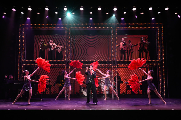 A performer sings centre stage in Return to Grace at Drayton Entertainment, surrounded by four dancers in sparkling costumes waving large red feather fans. Musicians perform behind them while additional singers appear on elevated platforms framed by illuminated marquee-style set pieces.