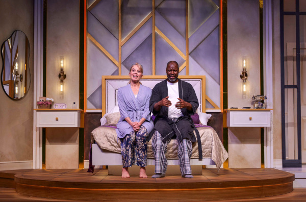 Two performers sit closely together on the edge of a bed, smiling broadly during a lighthearted moment. Both are dressed in sleepwear, with the bedroom’s geometric gold-trimmed wall panels and matching bedside tables visible behind them.