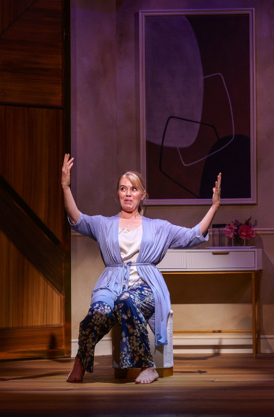 A performer sits barefoot on a stool in a lavender robe and floral pyjama pants, stretching both arms dramatically overhead while making an animated facial expression. A modern bedroom interior with wood panelling, abstract artwork, and a small console table fills the background.