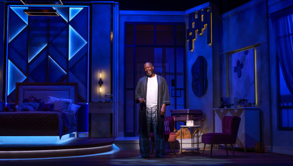 On a bedroom set washed in deep blue nighttime lighting, one performer stands centre stage in a dark robe and plaid pyjama pants with an expressive smile, while another lies asleep in the bed at the side of the stage. Chairs, a small table, and a bar area complete the stylish hotel-room setting.