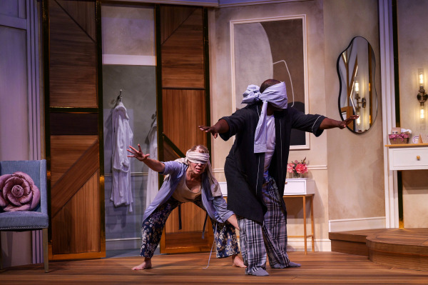 Two blindfolded performers cautiously make their way across the bedroom set with their arms stretched out in front of them. One bends low while reaching forward, while the other stands behind with a piece of fabric tied dramatically around their head and eyes.