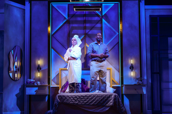 Under dramatic blue lighting, two performers stand on the bed in a tense moment. One wears a white bathrobe with a towel wrapped around their head, while the other stands beside them in a blue shirt and beige pants with hands clasped together.