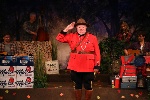 A comedic tableau unfolds in Sorry... I’m Not Sorry as a performer dressed in the iconic red uniform of a Royal Canadian Mounted Police officer stands centre stage delivering a crisp salute. Wearing the traditional red serge jacket, wide-brimmed hat, riding boots, and ceremonial belt, the performer strikes a proud pose while musicians playing guitar, fiddle, and upright bass accompany the scene from behind. The woodland-themed set is filled with camping gear, coolers, autumn leaves, stacked cases of Molson Canadian and Labatt Blue, and a sign reading “Keep Calm, eh?”, creating a playful and distinctly Canadian atmosphere that celebrates one of the country’s most recognizable national symbols.