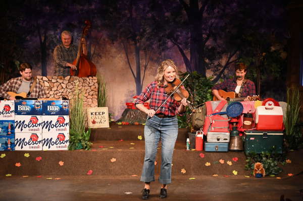 A lively musical performance unfolds in Sorry... I’m Not Sorry as a smiling performer in a red plaid shirt and rolled-cuff jeans plays the fiddle at centre stage. Behind them, members of the live band accompany the tune on guitar and upright bass, creating the feel of an energetic campfire jam session. The woodland-themed set is decorated with autumn leaves, camping gear, coolers, life jackets, and stacked cases of Canadian beer, while a sign reading “Keep Calm, eh?” reinforces the show’s playful celebration of Canadian culture. Warm lighting and the performer’s joyful expression contribute to the upbeat, community-focused atmosphere.