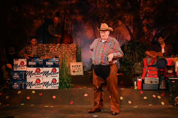 A humorous performance moment unfolds in Sorry... I’m Not Sorry as a performer dressed as a cowboy stands centre stage in a tan cowboy hat, plaid shirt, and oversized brown chaps. Striking a playful pose with one hand on a hip, the performer appears to be delivering a comedic monologue while musicians playing guitar, fiddle, and upright bass accompany the scene from behind. The woodland-themed set is decorated with camping gear, coolers, stacked cases of Molson Canadian and Labatt Blue, autumn leaves, and a sign reading “Keep Calm, eh?”, creating a lighthearted and distinctly Canadian atmosphere that celebrates rural stereotypes with warmth and humour.