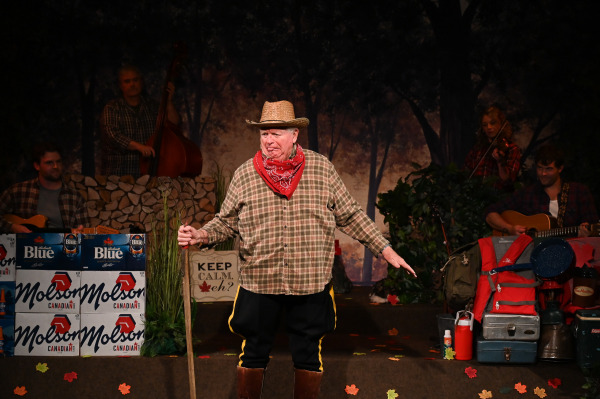 A playful character moment unfolds in Sorry... I’m Not Sorry as a performer dressed in a straw cowboy hat, red bandana, plaid shirt, and tall brown boots stands centre stage holding a long stick. With a mischievous expression and exaggerated pose, the performer embodies a humorous rural stereotype while a live band featuring guitar, fiddle, and upright bass accompanies the action from the background. The woodland-themed set is filled with camping supplies, coolers, autumn leaves, stacked cases of Molson Canadian and Labatt Blue, and a sign reading “Keep Calm, eh?”, creating a cheerful and distinctly Canadian atmosphere.