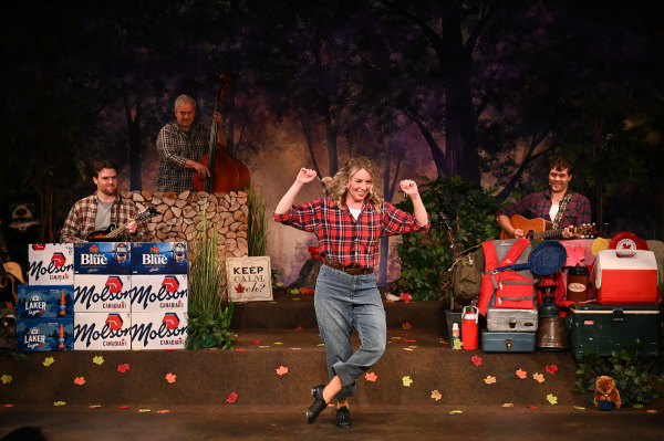 An energetic musical performance unfolds in Sorry... I’m Not Sorry as a performer dances joyfully at centre stage, smiling broadly while raising both arms and crossing one foot in front of the other. Dressed in a red plaid shirt, rolled blue jeans, and black shoes, the performer is framed by musicians playing mandolin, upright bass, and guitar against a woodland backdrop. The stage is decorated with camping gear, coolers, autumn leaves, stacked cases of Molson Canadian and Labatt Blue, and a sign reading “Keep Calm, eh?”, creating a playful and distinctly Canadian atmosphere. The warm lighting and lively movement capture the show’s celebratory humour and musical energy.
