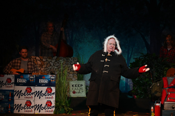 A humorous costume transformation takes centre stage in Sorry... I’m Not Sorry as a performer stands with arms outstretched, wearing an oversized black winter coat with toggle fastenings, red maple leaf mittens, and a white fur-trimmed hood framing their face. Behind them, members of the live band accompany the scene on guitar, fiddle, and upright bass. The woodland-inspired set features stacked cases of Canadian beer, camping gear, autumn leaves, and a sign reading “Keep Calm, eh?”, while cool blue lighting evokes a chilly northern landscape and enhances the playful celebration of Canadian stereotypes.