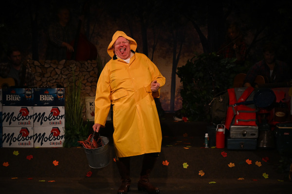 A comic performance moment unfolds in Sorry... I’m Not Sorry as a performer strides across the stage in a bright yellow rain slicker with the hood pulled up, carrying a metal bucket and making an exaggerated face with their tongue sticking out. The performer’s playful pose evokes a lighthearted parody of Canadian weather and outdoor life. Behind them, members of the live band are partially visible among a woodland-themed set decorated with autumn leaves, camping equipment, coolers, and stacked cases of Canadian beer. Warm stage lighting highlights the performer against the darker forest backdrop, emphasizing the show's energetic and distinctly Canadian humour.