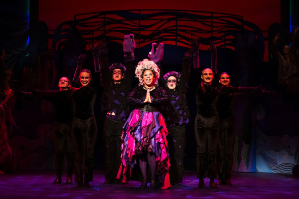 In a dramatic scene from Drayton Entertainment’s Disney’s The Little Mermaid at Hamilton Family Theatre Cambridge, a performer in a towering white wig and layered purple and pink costume stands centre stage with hands clasped while an ensemble dressed in dark, tattered costumes surrounds them with arms raised. Deep red and purple lighting and sweeping wave-like scenery create an ominous underwater setting.