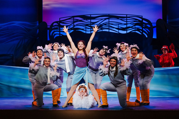 In a colourful musical number from Drayton Entertainment’s Disney’s The Little Mermaid at Hamilton Family Theatre Cambridge, Ariel stands centre stage with both arms raised as an ensemble of seabird performers in grey costumes, feathered trim, goggles, and orange leg coverings surrounds her in energetic poses. A performer dressed in bright red watches from the background against a vivid blue underwater set.