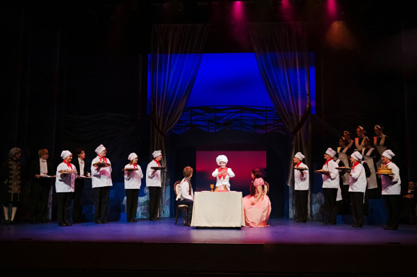 : In a grand dining scene from Drayton Entertainment’s Disney’s The Little Mermaid at Hamilton Family Theatre Cambridge, Ariel and a young performer sit opposite each other at a small table while a chef stands between them presenting the meal. An ensemble of chefs holding trays lines both sides of the stage, with additional attendants gathered on raised platforms beneath sweeping curtains and colourful theatrical lighting.
