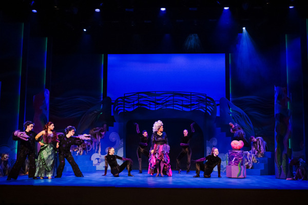 In a dramatic underwater scene from Drayton Entertainment’s Disney’s The Little Mermaid at Hamilton Family Theatre Cambridge, Ariel stands at one side of the stage surrounded by dark-costumed sea creatures while a performer in a voluminous purple-and-pink costume stands centre stage. Additional ensemble members crouch and pose across the set beneath deep blue lighting, with wave-shaped scenery, staircases, and a glowing shell-like prop completing the ominous underwater setting.