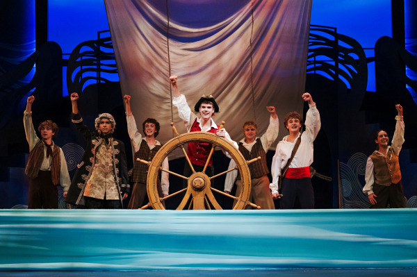 In a shipboard scene from Drayton Entertainment’s Disney’s The Little Mermaid at Hamilton Family Theatre Cambridge, a performer in a red vest and black tricorn hat stands behind a large wooden ship’s wheel with one fist raised. An ensemble of sailors in period-inspired costumes surrounds the wheel with arms lifted beneath a large sail, while stylized blue waves stretch across the front of the stage.
