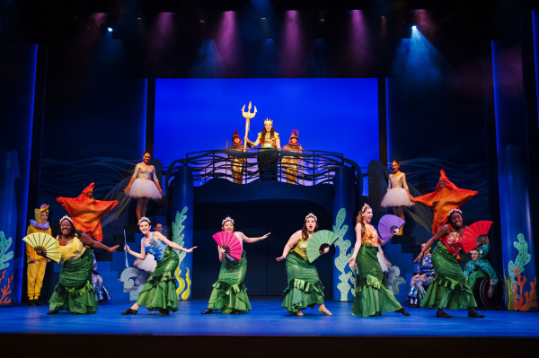 In a vibrant underwater ensemble scene from Drayton Entertainment’s Disney’s The Little Mermaid at Hamilton Family Theatre Cambridge, performers in shimmering green mermaid costumes dance across the front of the stage holding colourful folding fans. Above them, a crowned performer holding a golden trident stands on a raised platform surrounded by fantastical sea creatures, including starfish and other brightly costumed marine characters.