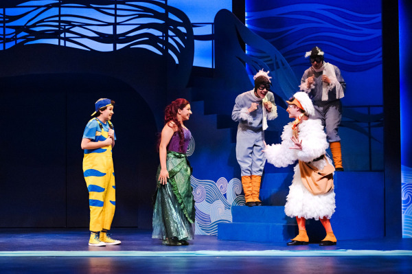 In a colourful scene from Drayton Entertainment’s Disney’s The Little Mermaid at Hamilton Family Theatre Cambridge, a performer in a shimmering green mermaid costume smiles toward a performer in an elaborate white seabird costume, who gestures animatedly with both hands. A performer in bright blue and yellow watches from the side while two additional seabird performers perch on blue steps behind them amid stylized wave scenery.