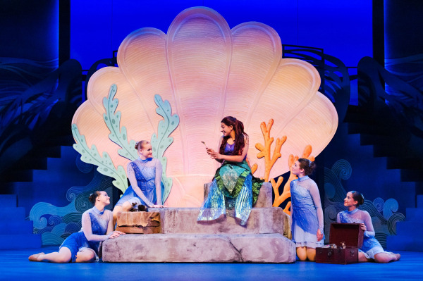 In an underwater scene from Drayton Entertainment’s Disney’s The Little Mermaid at Hamilton Family Theatre Cambridge, Ariel sits on a rocky platform in front of an enormous glowing seashell while four performers in sparkling blue costumes gather around her. Ariel holds up a small object as the others watch, with colourful coral pieces and stylized waves completing the grotto-like setting.