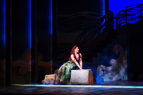 In a scene from Drayton Entertainment’s Disney’s The Little Mermaid at Hamilton Family Theatre Cambridge, Ariel sits alone on a rocky platform, smiling as she looks offstage. Her long red hair and shimmering purple-and-green mermaid costume stand out against the dark blue underwater setting, framed by stylized waves, tall vertical scenic panels, and a staircase rising behind her.