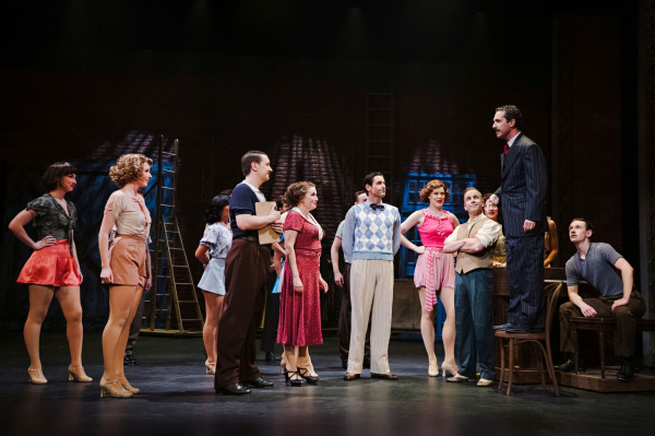 A group of performers in 1930s-inspired rehearsal attire gathers around a man standing on a chair in 42nd Street. The ensemble watches attentively as he addresses them, creating the atmosphere of a Broadway audition or rehearsal. The industrial backstage set features brick walls, ladders, and stage equipment, while warm theatrical lighting focuses attention on the spirited group dynamic.