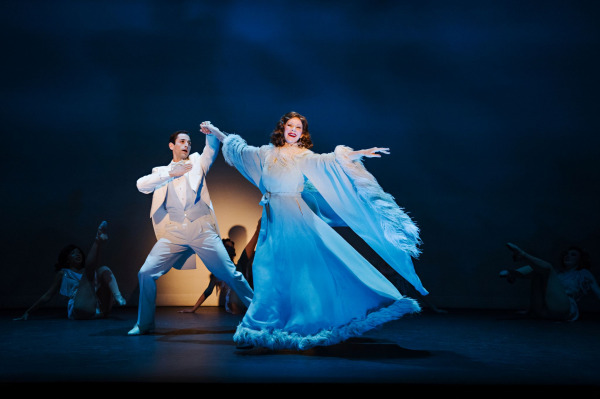 Two performers dance beneath cool blue lighting in a glamorous production number from 42nd Street. One wears a flowing pale-blue gown trimmed with feathers, while the other is dressed in a crisp white tuxedo. Their elegant poses are framed by shadowy dancers in the background, creating a dreamlike, old-Hollywood atmosphere.
