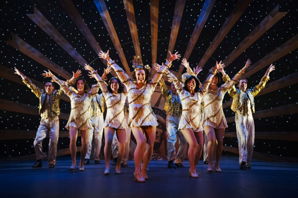 A chorus line of performers in sparkling gold costumes advances toward the audience in a high-energy dance number from 42nd Street. With arms raised and bright smiles, the ensemble performs beneath a starry backdrop and radiating Art Deco set pieces, capturing the spectacle and precision of a classic Broadway production.