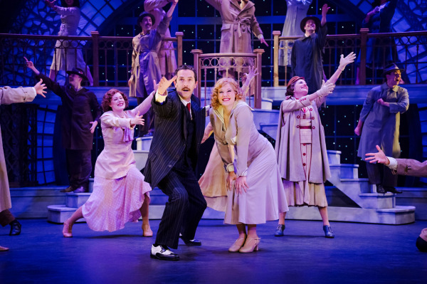 Two lead performers pose centre stage during a large ensemble number in 42nd Street. Surrounded by dancers dressed in period coats and dresses, the pair smiles toward the audience while grand staircase scenery and glowing star-like lights create a romantic, theatrical backdrop.