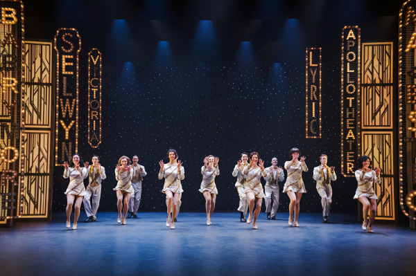 The ensemble of 42nd Street performs a synchronized tap dance routine beneath towering illuminated Broadway marquee signs. Dressed in coordinated white and gold costumes, the performers advance across the stage in perfect formation, showcasing the precision choreography and dazzling spectacle of the production.