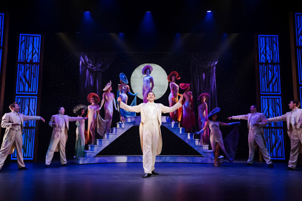 A performer in a white tailcoat stands centre stage with arms outstretched during a lavish musical sequence in 42nd Street. Ensemble members in shimmering gowns and wide-brimmed hats pose atop symmetrical staircases beneath a glowing full moon backdrop. Art Deco-inspired scenery and dramatic lighting evoke the glamour of Broadway’s Golden Age.