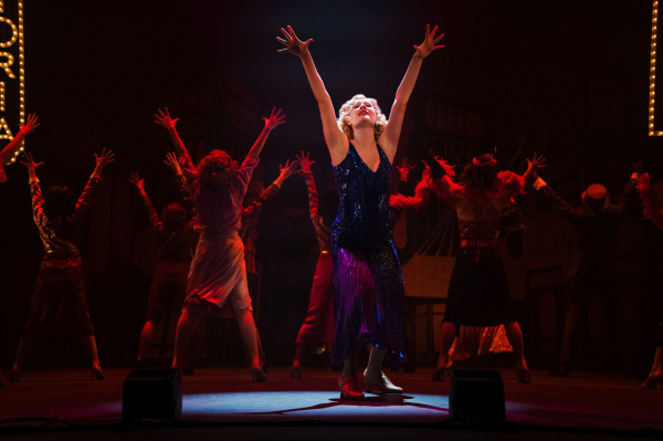 A performer in a sparkling blue dress stands in a spotlight during a dramatic musical number from 42nd Street. Bathed in rich red lighting, they reach both arms upward while ensemble members pose behind them with matching gestures. Marquee-style Broadway signs and cityscape scenery frame the powerful performance.