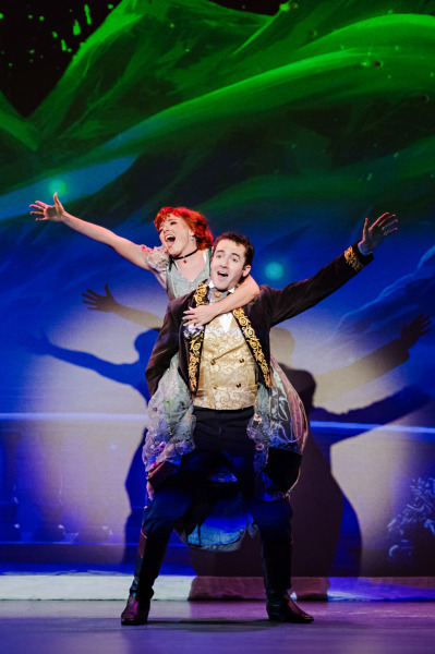 Two performers share an exuberant moment onstage, with one riding piggyback on the other as both stretch their arms outward. They wear richly detailed period costumes against a vivid blue and green projected backdrop.