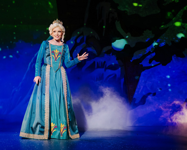 A performer wearing a jeweled crown and floor-length teal gown stands in a snowy forest setting, gesturing with one hand. Deep blue and green lighting, silhouetted trees, scattered snow, and low-lying mist create a wintry atmosphere.